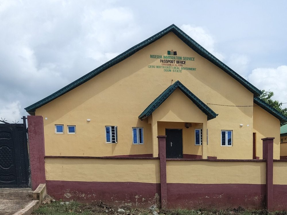 A yellow single-storey Nigeria Immigration Service passport office with a green roof, behind a low wall