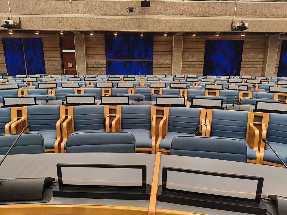Rows of empty blue delegate chairs with microphones in a conference hall