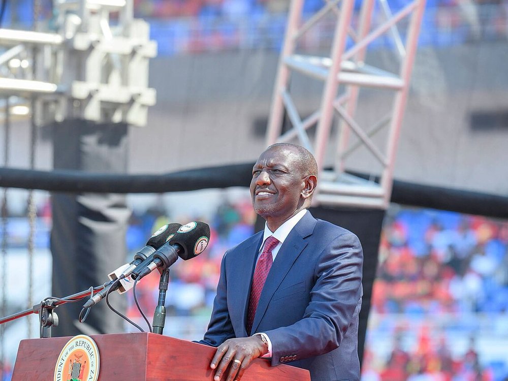 President William Ruto smiling at a podium with microphones, in front of a stadium crowd